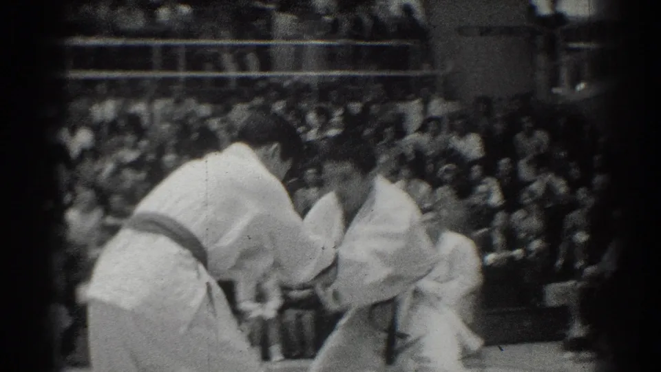 A close spectator view shows one karate competitor bending low as the opponent reaches in, with the crowd visible behind them.