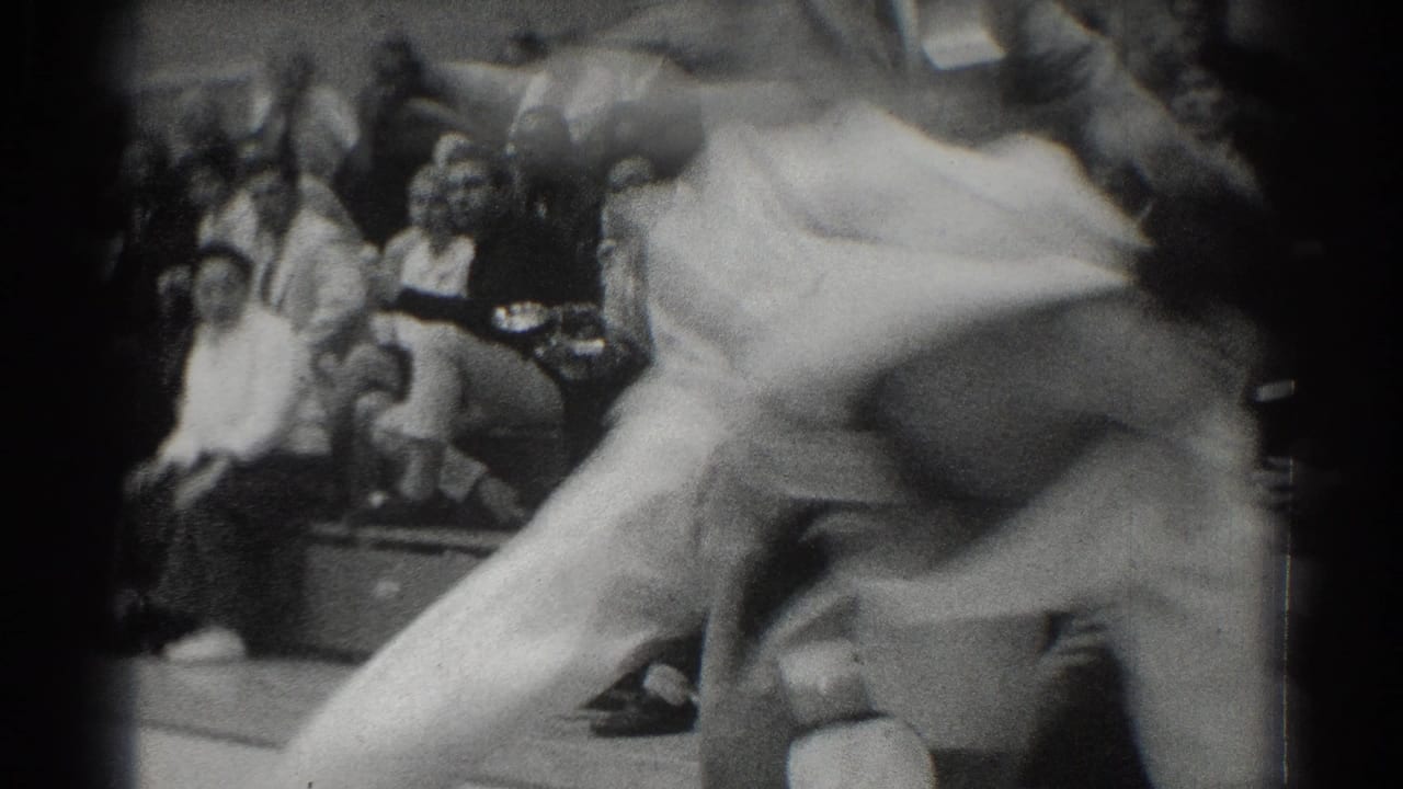 Two judo athletes in white gi grapple on a mat before seated spectators.