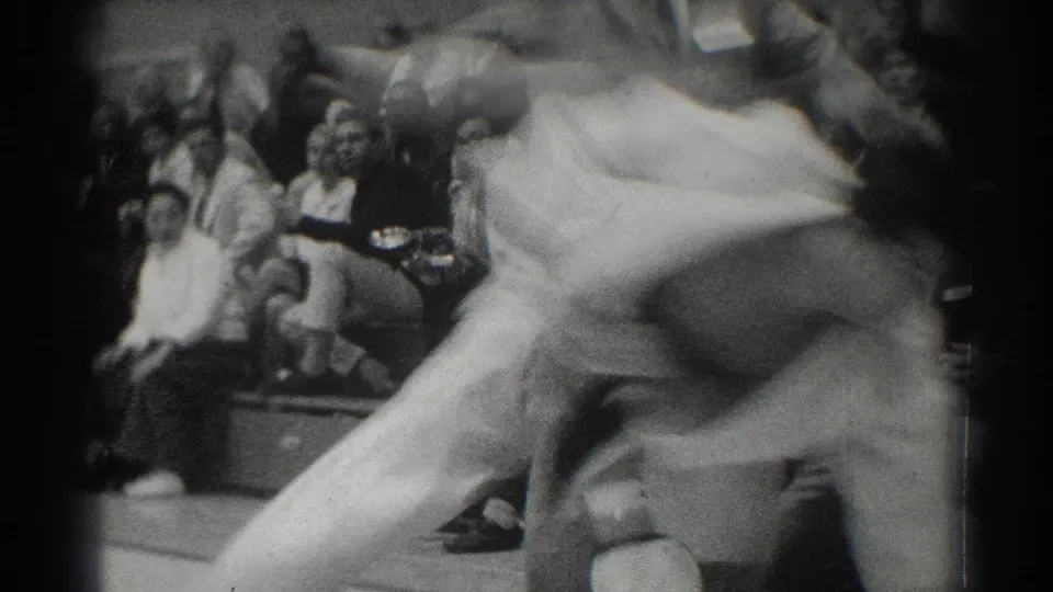 In 1966 West Covina footage, two judo competitors in white gi are caught mid-grapple near a seated audience.