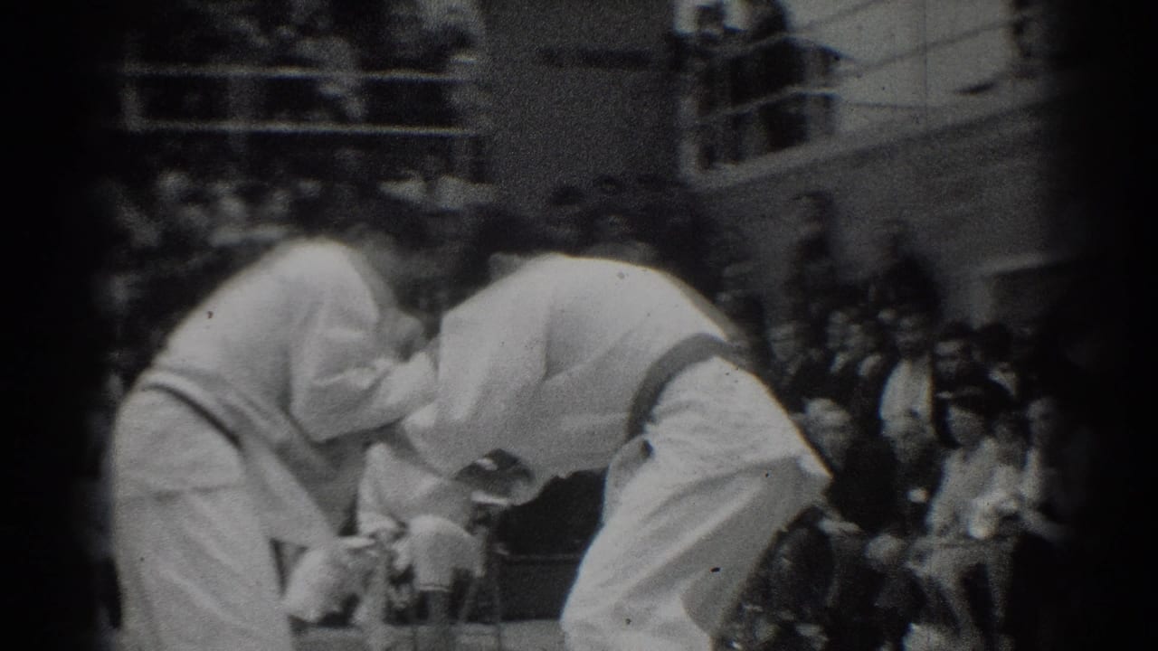Two judo competitors bow or lean toward each other on the mat as spectators watch.