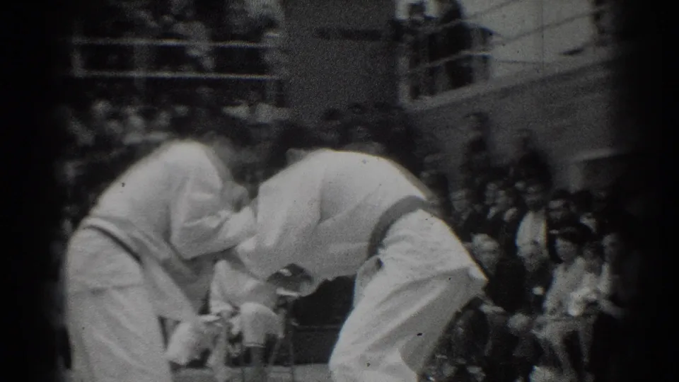 Two athletes in white gi face each other on the mat in the West Covina judo competition, with bleachers behind them.