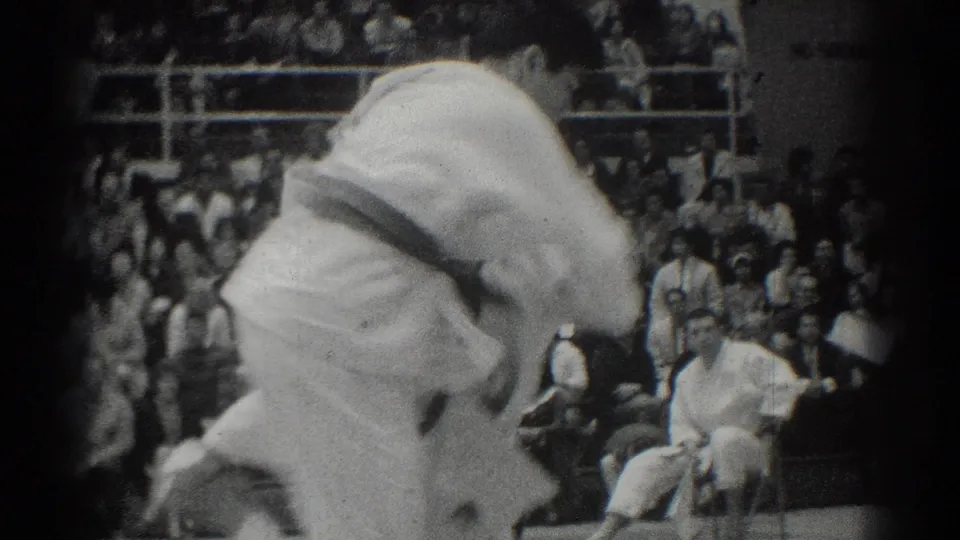 A white-gi competitor bends into a throw or hold during the 1966 judo match as spectators fill the background.