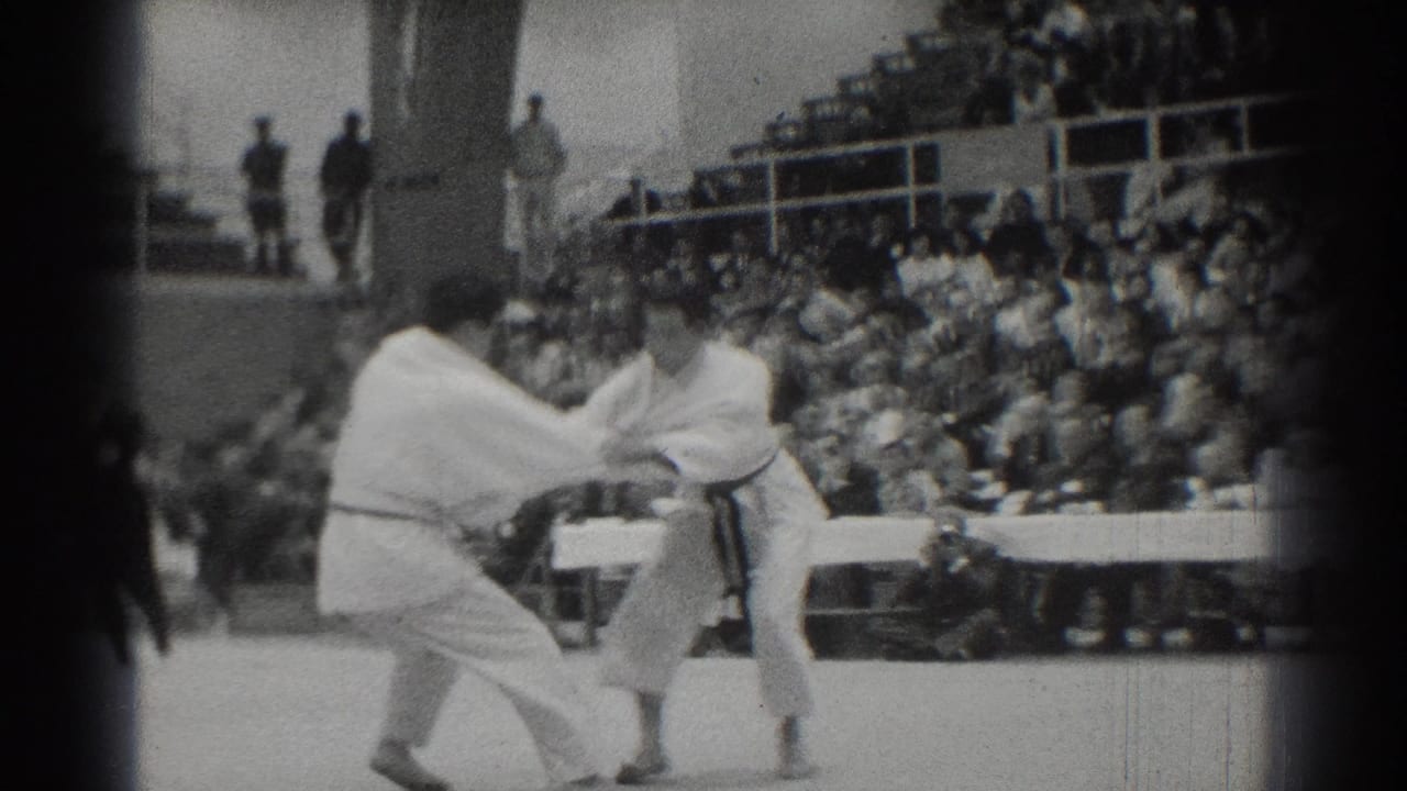 Two men in white gi grapple on a mat in front of crowded bleachers.