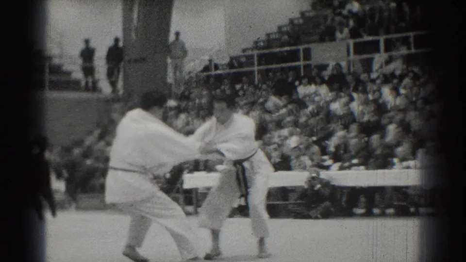 1966 West Covina 8mm frame showing two martial arts competitors in white gi grappling before spectators in bleachers.