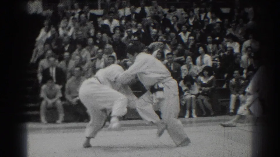 Martial arts competitors in white gi appear mid-throw before a dense seated crowd in West Covina, California, in 1966 footage.