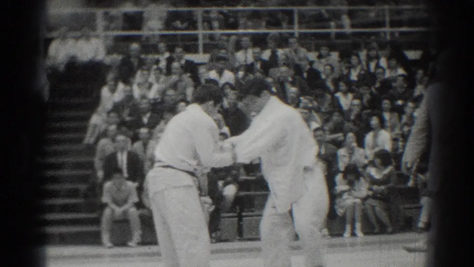Black-and-white 8mm frame of two martial arts competitors resetting or gripping on the mat before a seated audience.