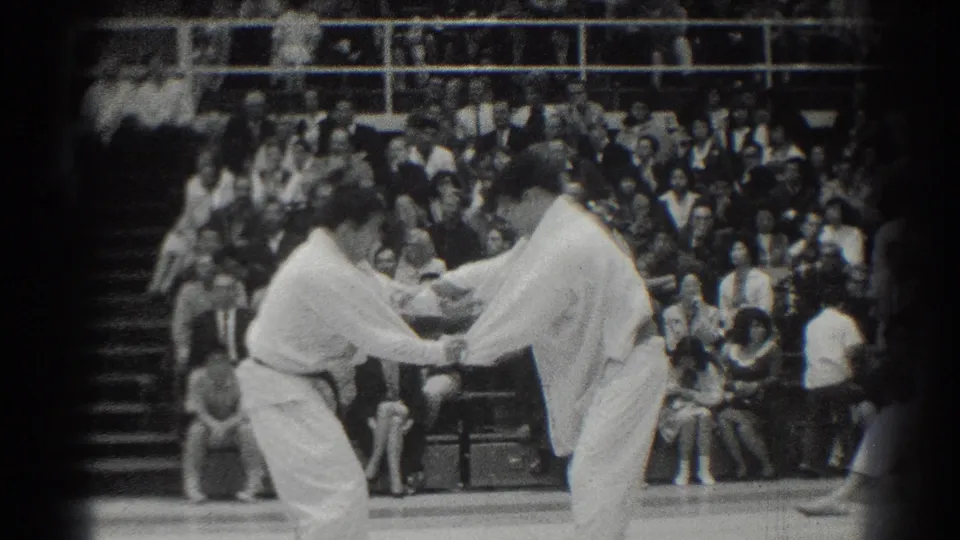 West Covina 1966 home-movie frame showing two martial arts competitors gripping on the mat with a suited official at the side.