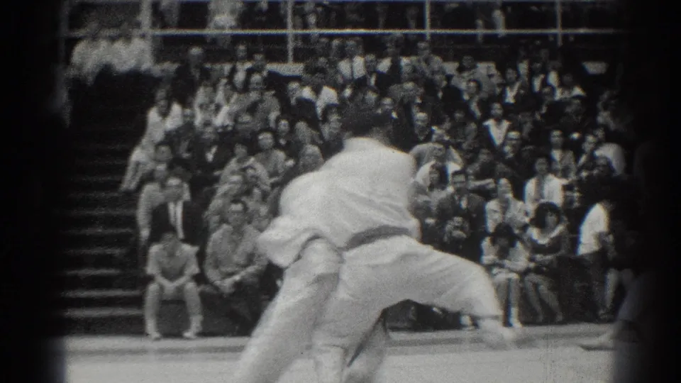 In the supplied 1966 West Covina context, two karate competitors in white gi grapple at close range while spectators fill the bleachers behind them.
