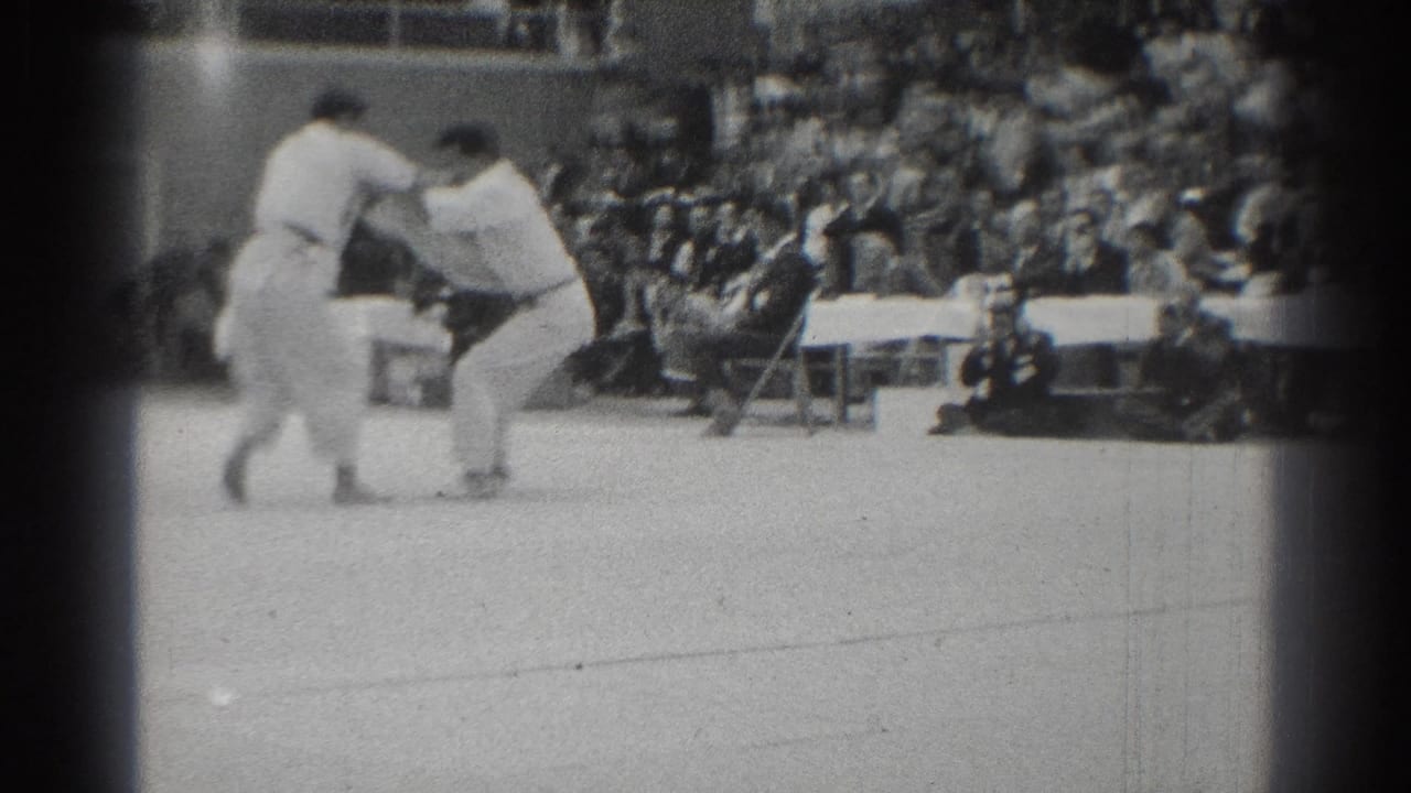 Two white-gi competitors appear to engage from a standing position during the karate match, with a seated audience and folding chairs visible along the edge of the floor.