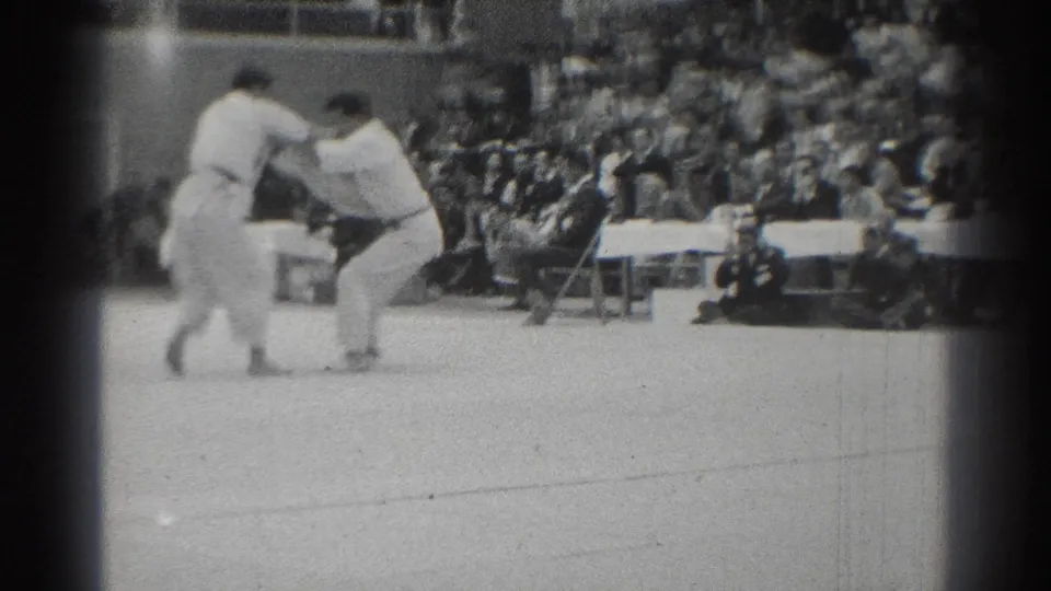 Two white-gi competitors appear to engage from a standing position during the karate match, with a seated audience and folding chairs visible along the edge of the floor.