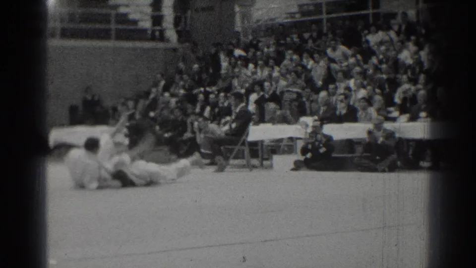 A competitor appears to land or slide on the gym floor during a takedown in the 1966 West Covina karate match, with the audience watching from bleachers behind.