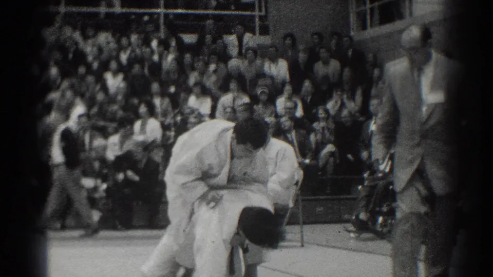 A close view shows one white-gi competitor bent over another during grappling action, while a suited referee observes beside the crowded gym audience.