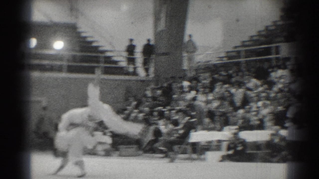 Two martial artists in white uniforms move on a gym mat before crowded bleachers.