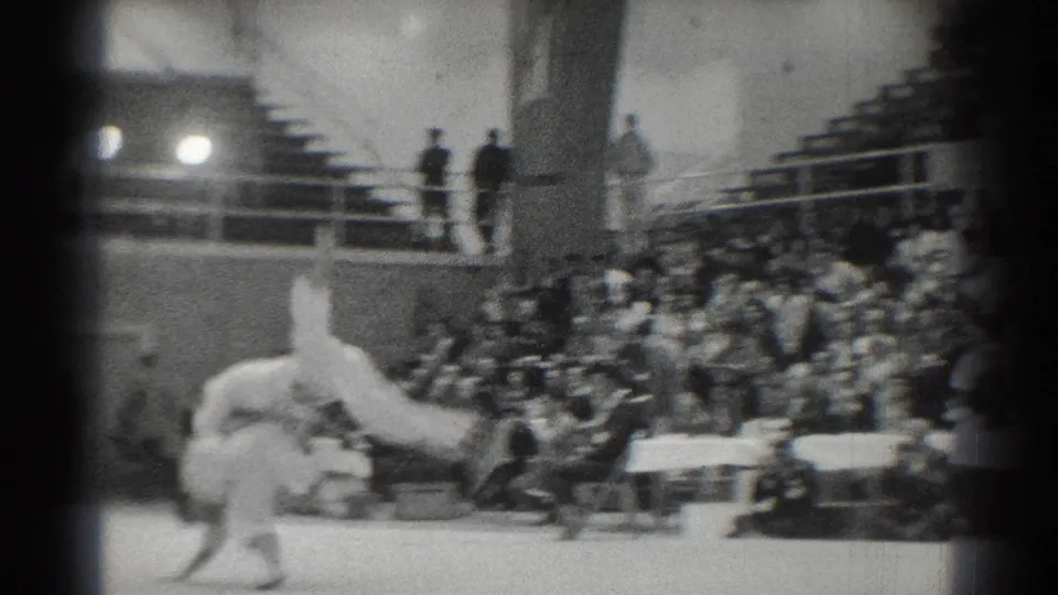 1966 West Covina home-movie frame showing two martial artists in white gi during an indoor gym match with spectators behind them.