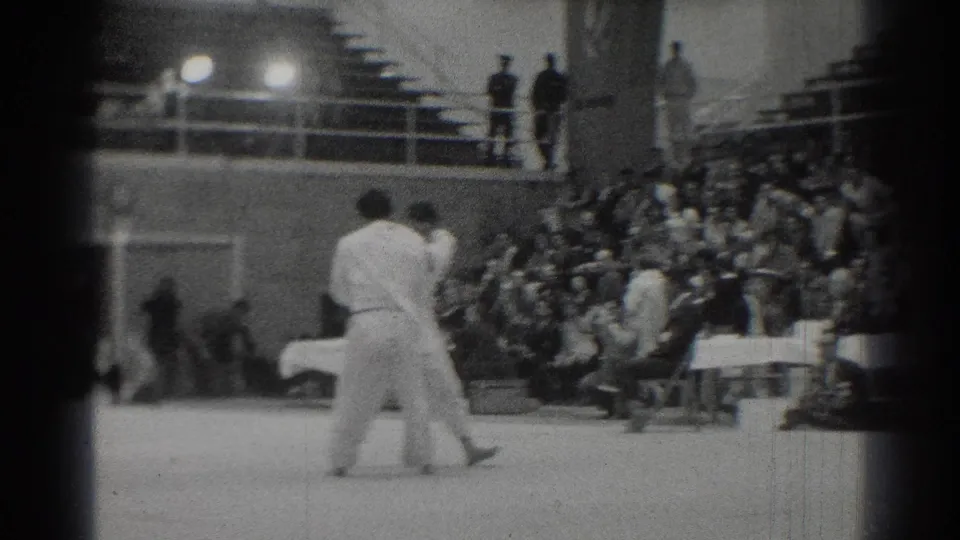Two competitors in white gi face each other on a gym mat during the 1966 West Covina martial arts event, with bleachers behind.