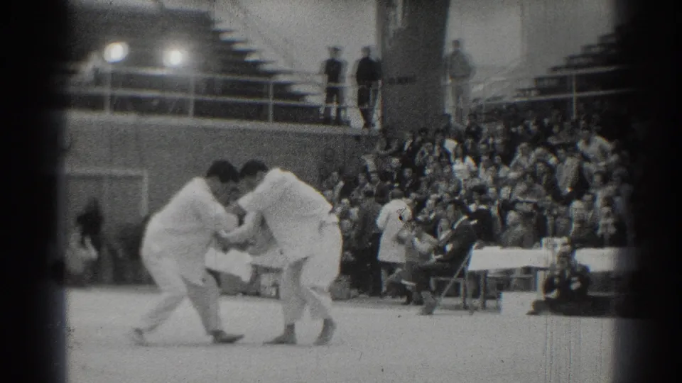 A close grappling moment in the 1966 West Covina gym match shows two white-uniformed martial artists with seated spectators behind them.
