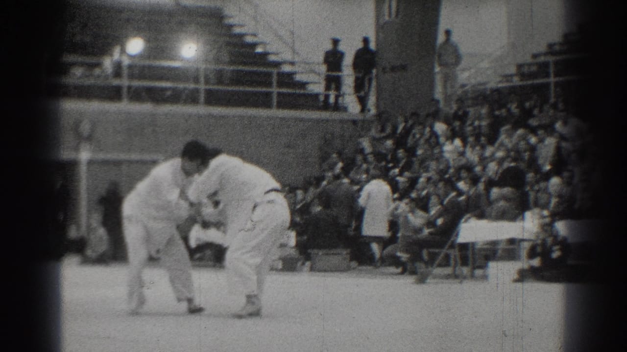 Two judo competitors crouch and grip on a mat before crowded bleachers.