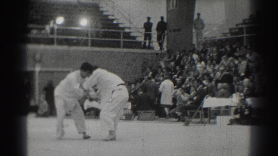 Two judo athletes in white judogi grapple on an indoor mat in 1966 West Covina, with spectators seated behind them.