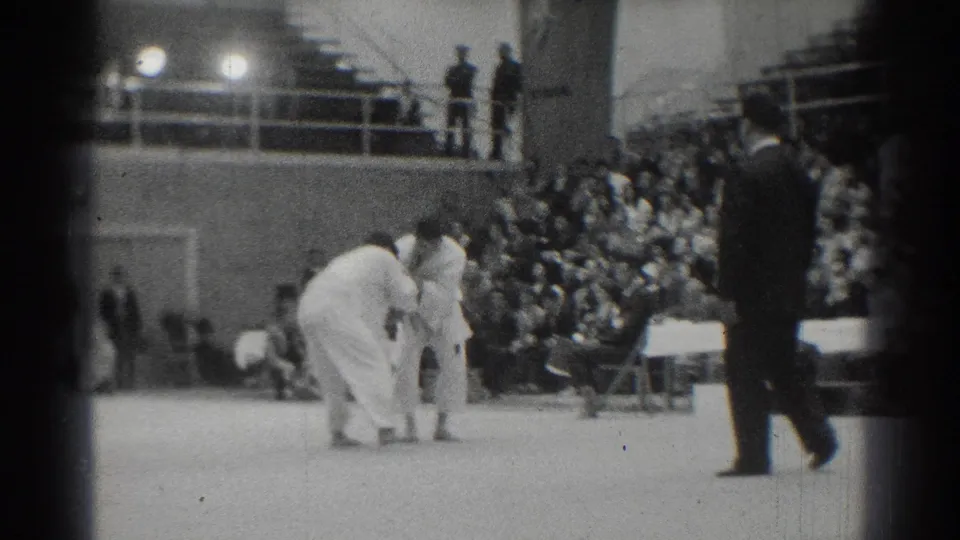 The athletes bend toward each other on the judo mat as a suited referee crosses the foreground in the 1966 indoor arena.