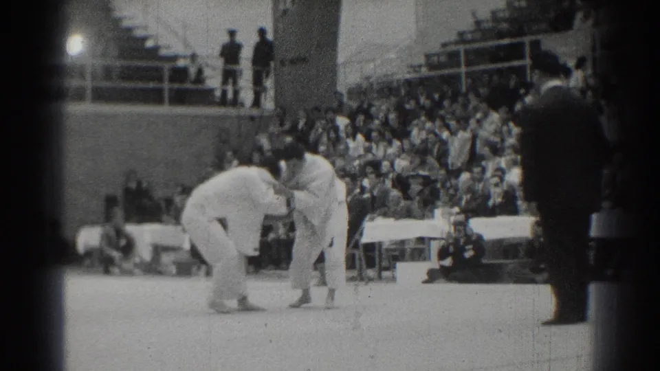 Two athletes continue grappling on the mat while the dark-suited referee observes beside the crowded West Covina arena seating.