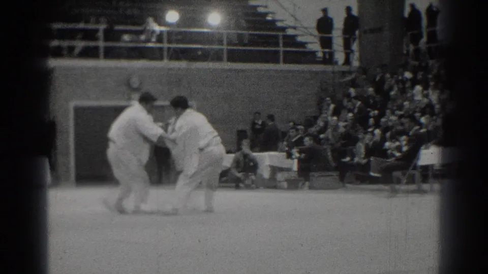 Two men in white judo gis square off on a gym mat in 1966 West Covina footage as spectators watch from the side.