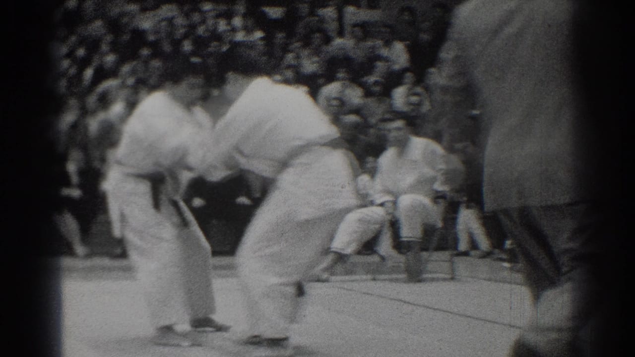 The competitors hold each other's gis during the match, with a crowded gym audience visible behind them in West Covina.