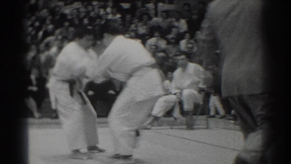 The competitors hold each other's gis during the match, with a crowded gym audience visible behind them in West Covina.