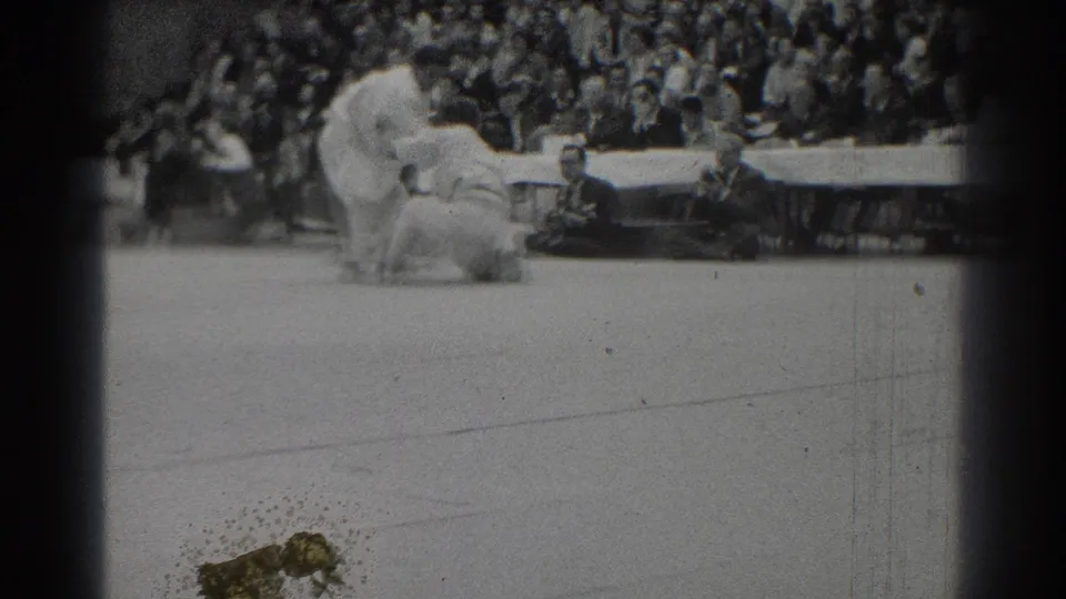 After the throw, one competitor lies on the mat while the other stands over him, with officials and spectators watching.