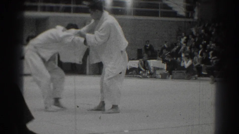1966 West Covina home-movie frame showing two judo athletes beginning a grip exchange on an indoor competition mat.