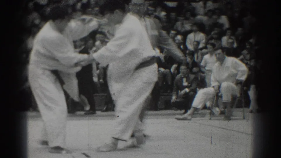 1966 West Covina frame with two judo athletes gripping at close range as a seated official and audience observe the match.