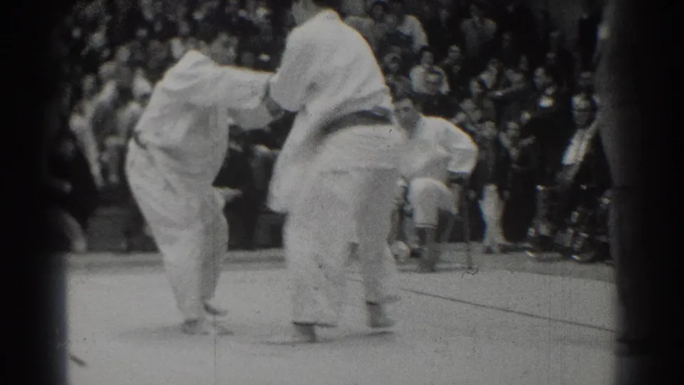 1966 West Covina home-movie frame showing a continuing judo grip exchange with spectators gathered closely behind the mat.