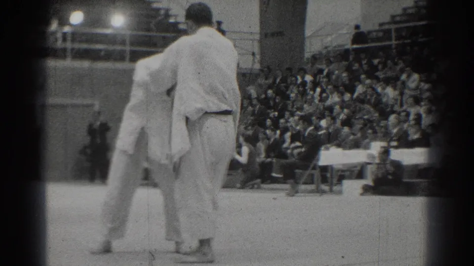 In 1966 West Covina footage, two adult competitors in white gi uniforms face each other on a mat with bleachers behind them.