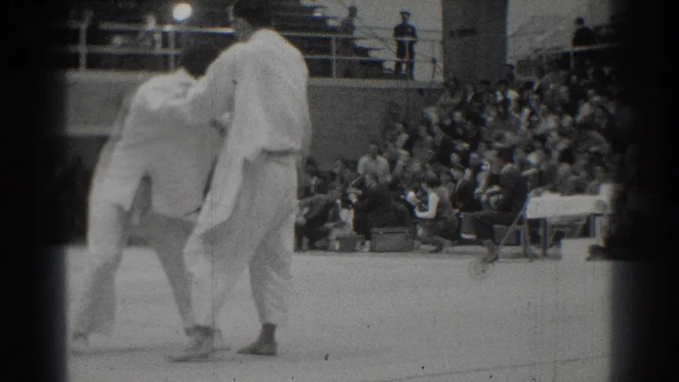 The West Covina martial arts match continues as two adult men in white gi uniforms shift position before the bleacher crowd.