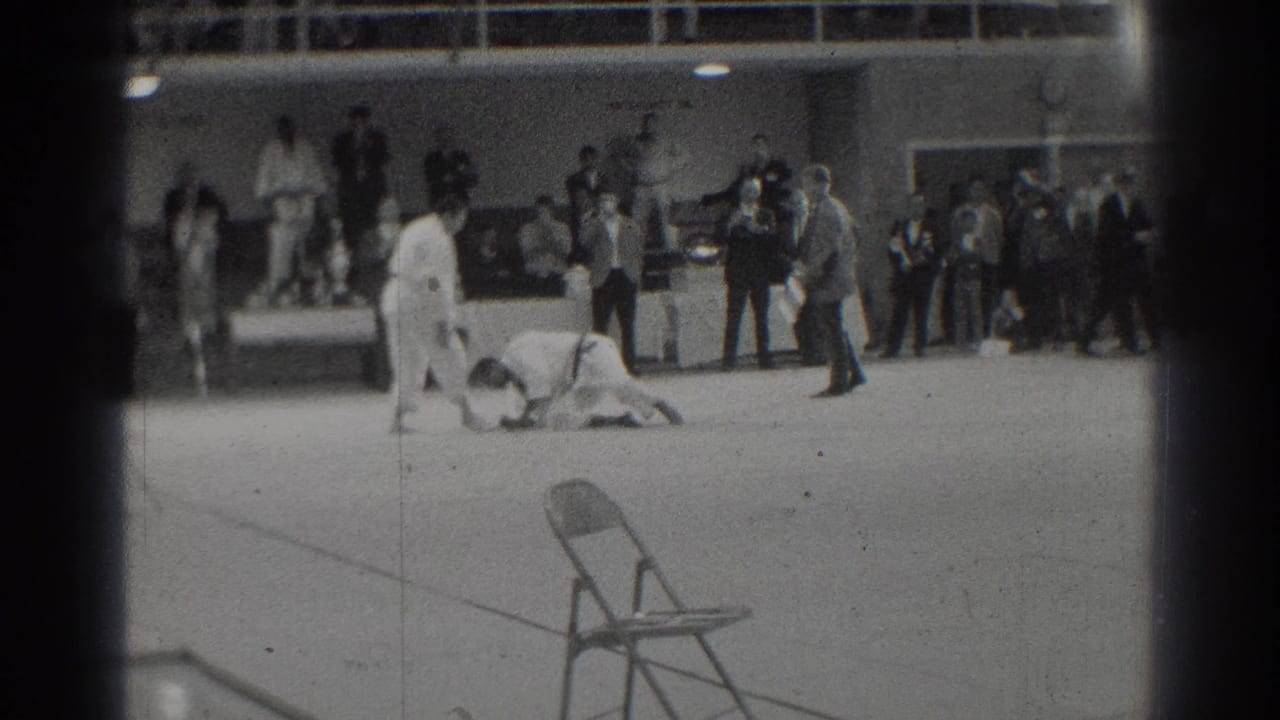 Two judo athletes grapple on a mat while spectators stand along the back wall.