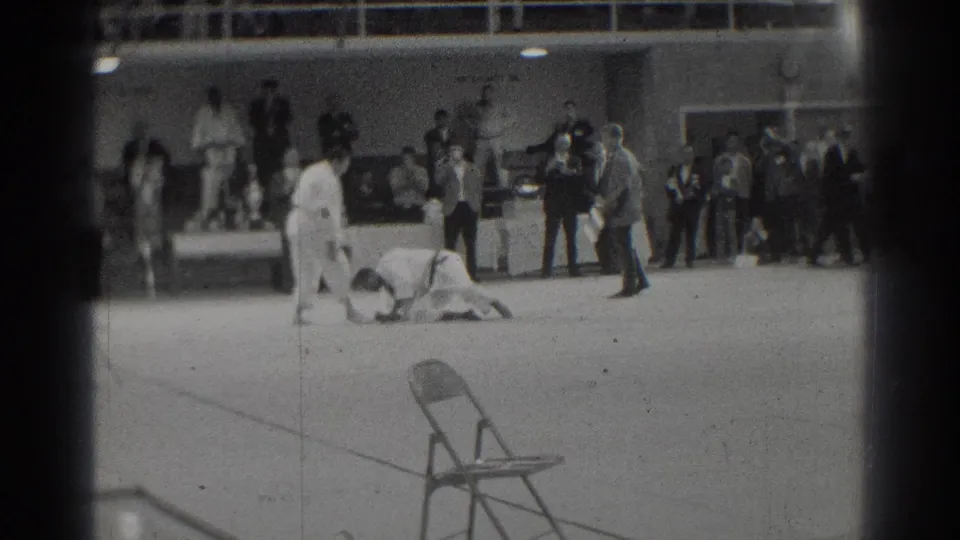 1966 West Covina judo match with athletes in white gi on the mat, spectators watching, and a folding chair in the foreground.