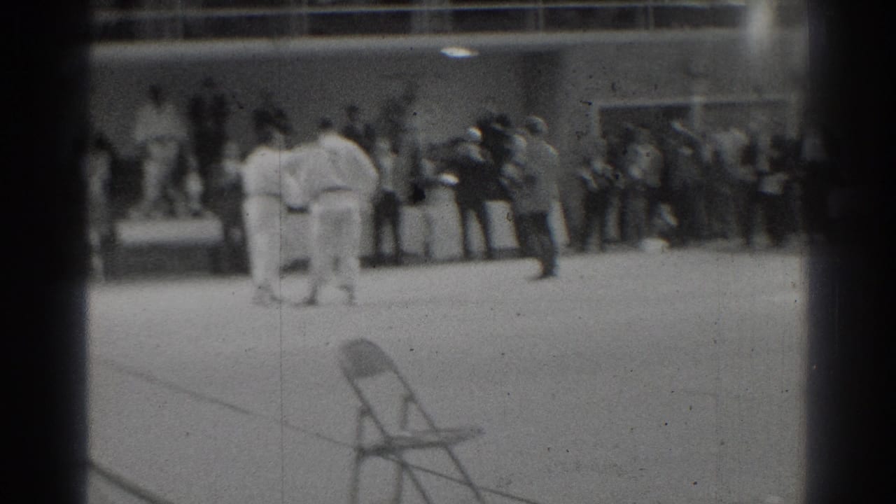 Two judo participants in white gi stand in a gym-like room as a group of spectators watches from the back wall.