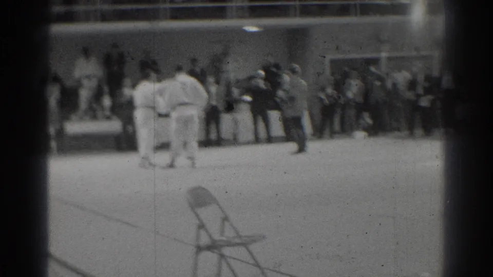 Two judo participants in white gi stand in a gym-like room as a group of spectators watches from the back wall.