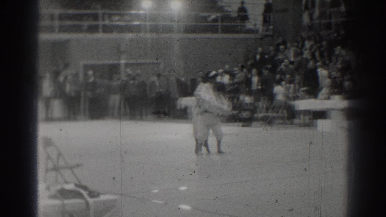 Two judo practitioners on a mat with spectators seated in bleachers behind them.