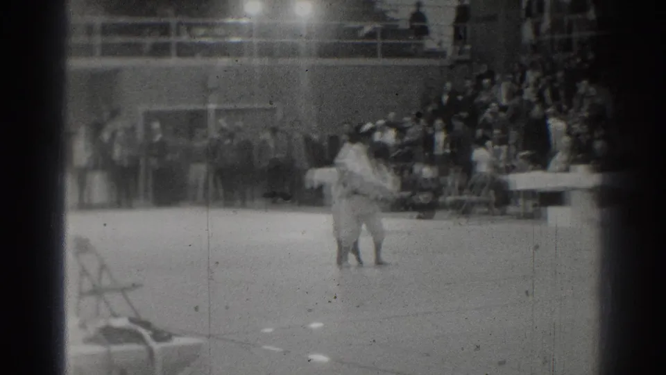 1966 West Covina home-movie frame showing two judo practitioners on a gym mat while spectators watch from bleachers.