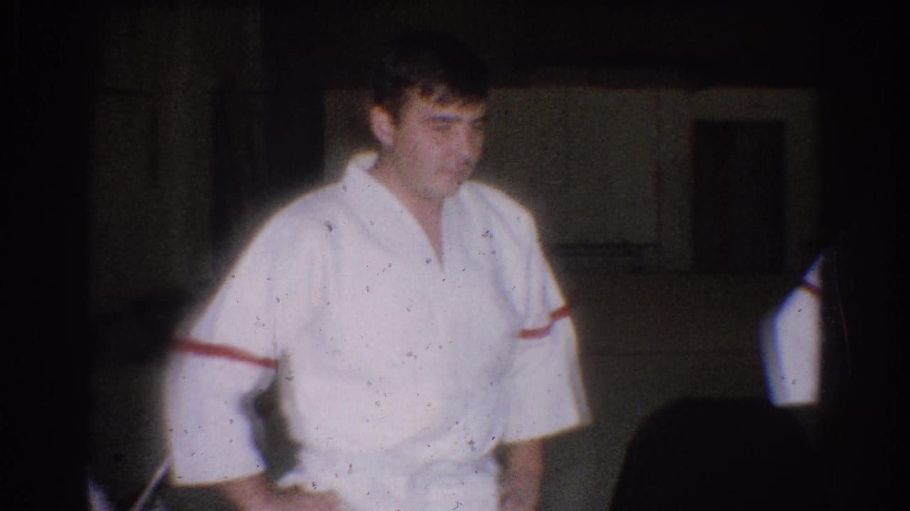 A judo practitioner in a white gi with red trim stands in a dim West Covina gym during the 1966 training footage.