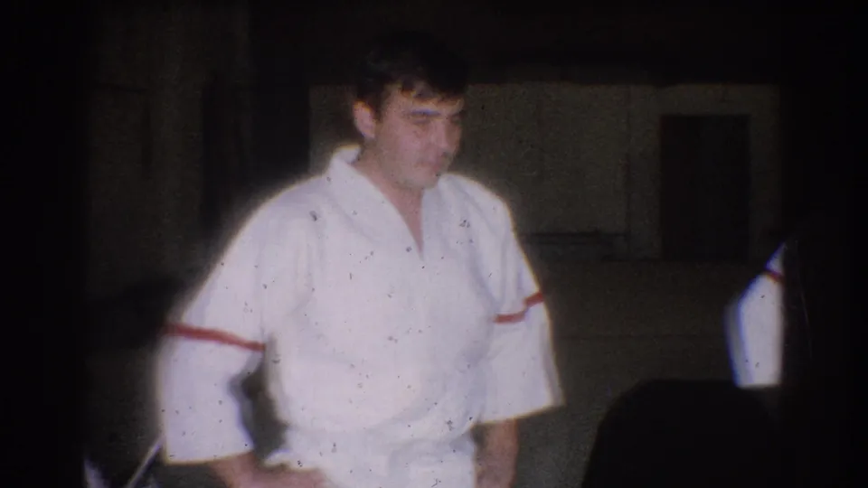 A judo practitioner in a white gi with red trim stands in a dim West Covina gym during the 1966 training footage.