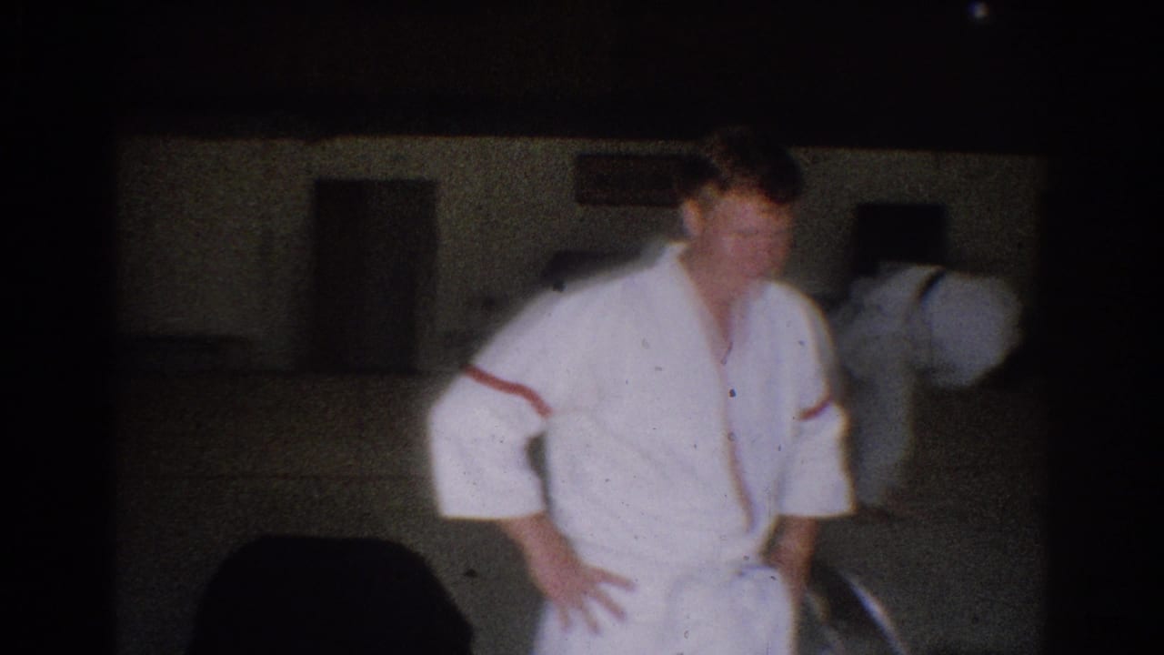 1966 West Covina gym frame with a judo practitioner standing on the mat while another participant bends in the background.