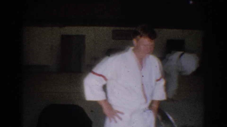 1966 West Covina gym frame with a judo practitioner standing on the mat while another participant bends in the background.