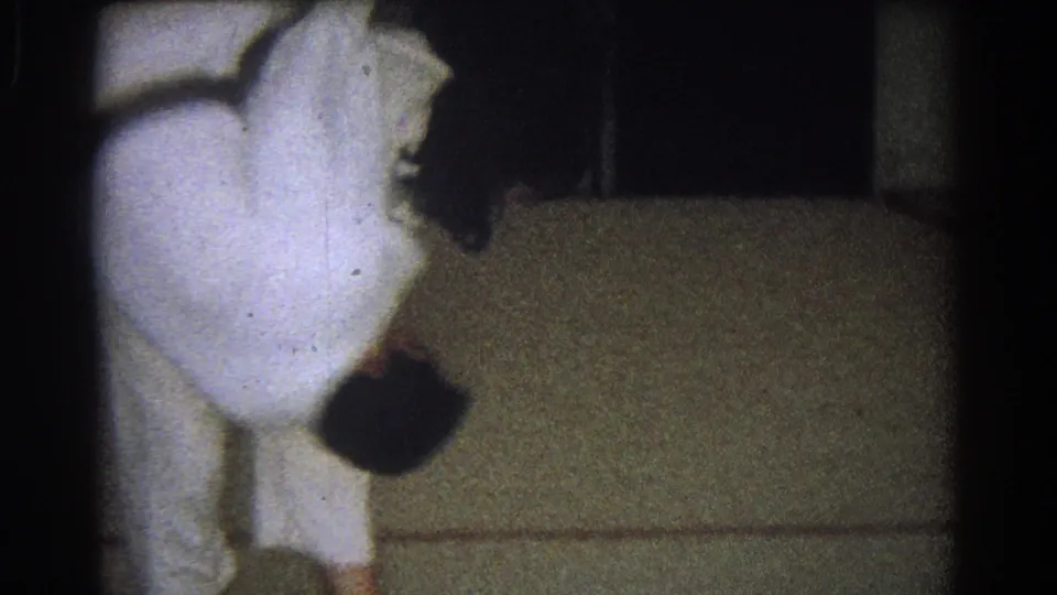 In 1966 West Covina home-movie footage, two adults in white martial arts uniforms begin a close-contact throw near an indoor mat.