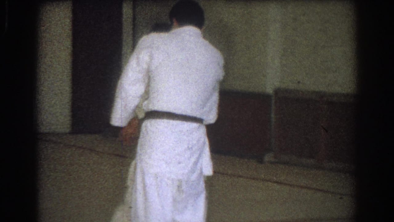 A practitioner in a white uniform bends forward during a throw or setup while another adult stands close in the indoor practice space.
