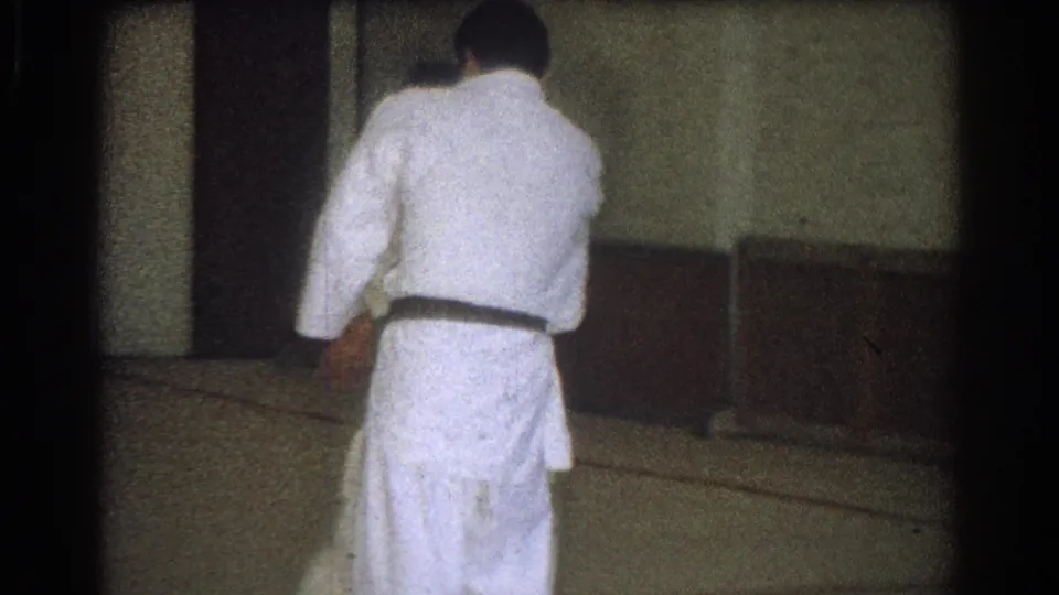 A practitioner in a white uniform bends forward during a throw or setup while another adult stands close in the indoor practice space.