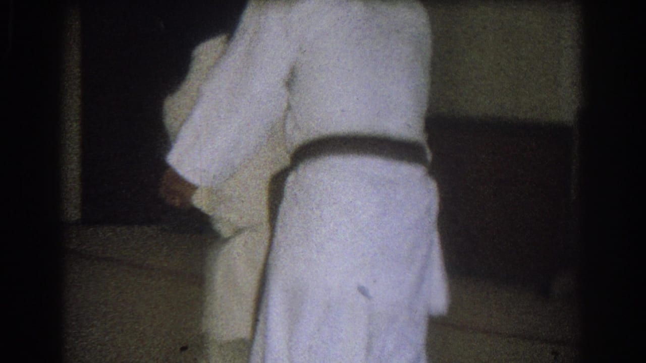Close 8mm view of two adults in white martial arts uniforms as one appears to execute or complete a throw in the indoor practice room.