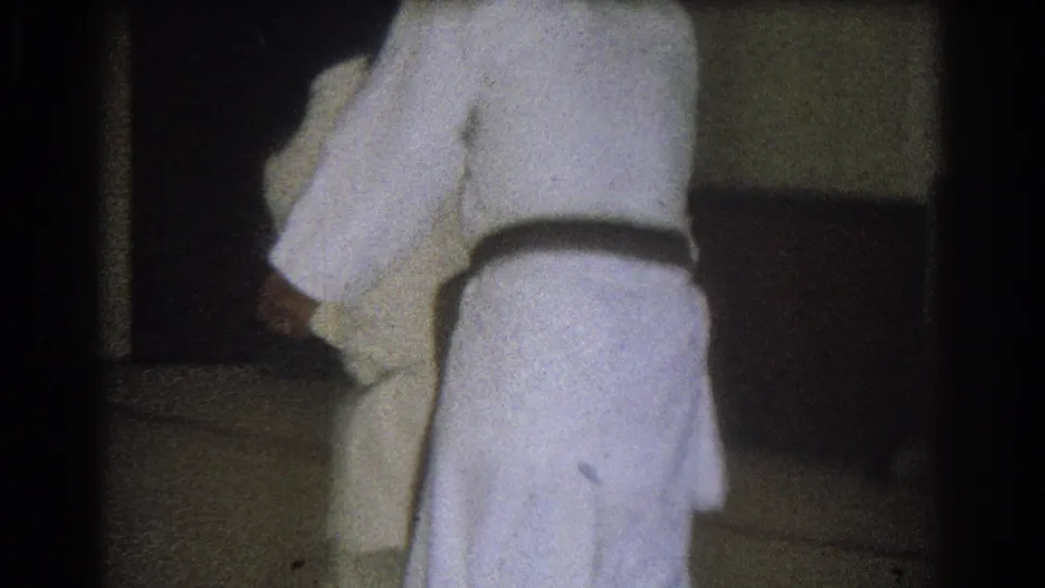 Close 8mm view of two adults in white martial arts uniforms as one appears to execute or complete a throw in the indoor practice room.