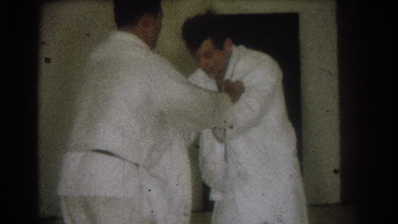 In 1966 West Covina home-movie footage, two men in white gi face each other while practicing a close grappling hold indoors.