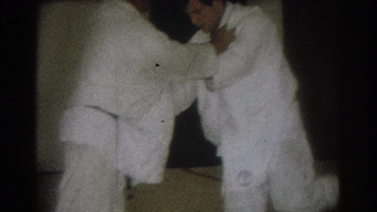 Two men in gi continue a standing grappling move in a dim room.